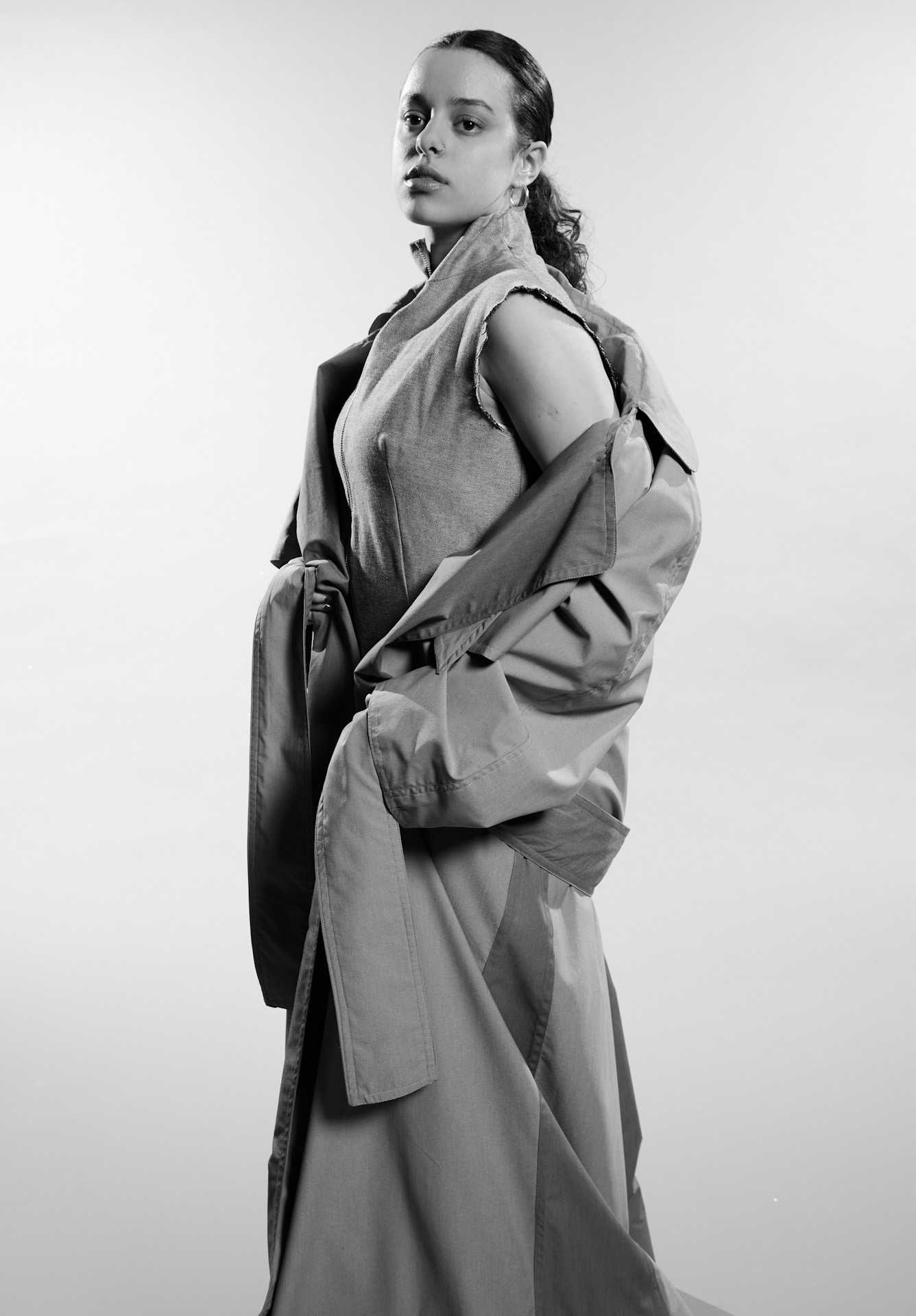 The model wears a full length trench coat with a knitted vest worn beneath it. The model clutches the belt of the coat as she looks beyond the camera lens.